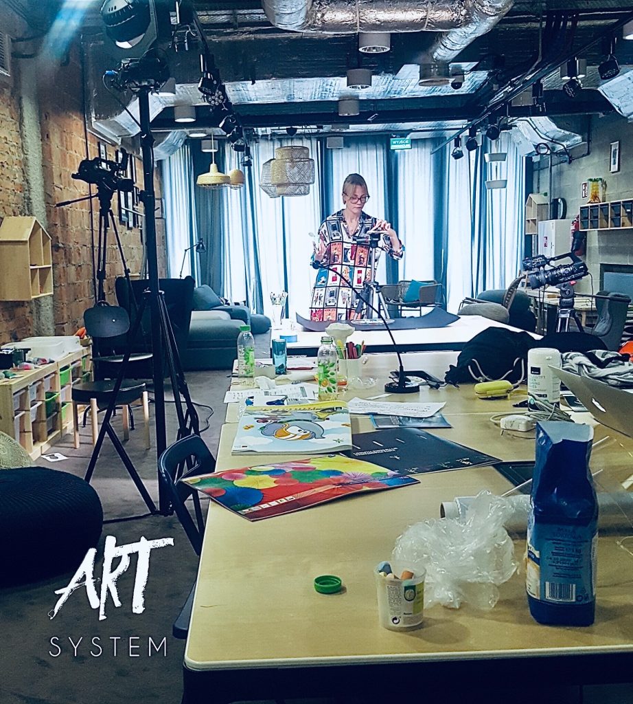 Widok wnętrza nowoczesnej przestrzeni warsztatowej CSK. Na pierwszym planie stół z materiałami plastycznymi, rysunkami i sprzętem do nagrywania. W tle kobieta w kolorowej sukience prowadzi zajęcia Poklatkowo przy statywie i kamerze. Na ścianie cegła, wokół lampy, kurtyny i przytulne meble.